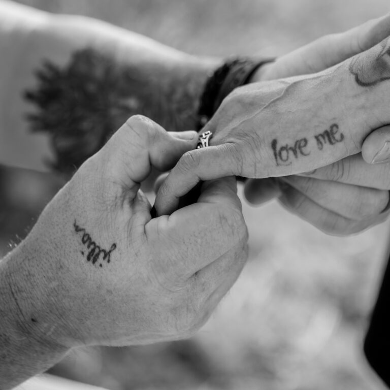 A black and white close-up photo of a couple gently holding hands. One hand has a tattoo that reads "love me," and the other is marked with the word "Yuma." A ring is visible on one finger, emphasizing the intimacy and connection.