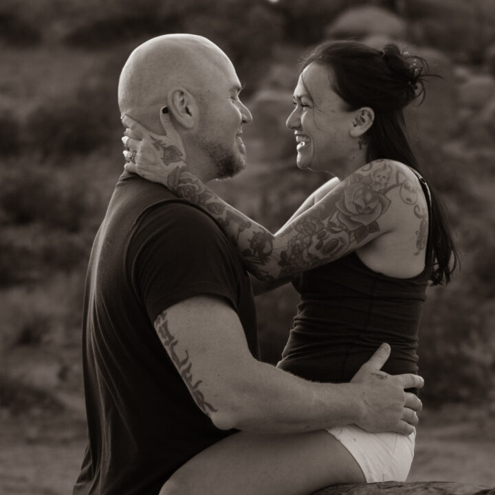 A sepia-toned photo of the couple sharing a playful, loving moment. The man sits on a rock while the woman, with tattooed arms, sits on his lap, both smiling at each other with carefree affection.
